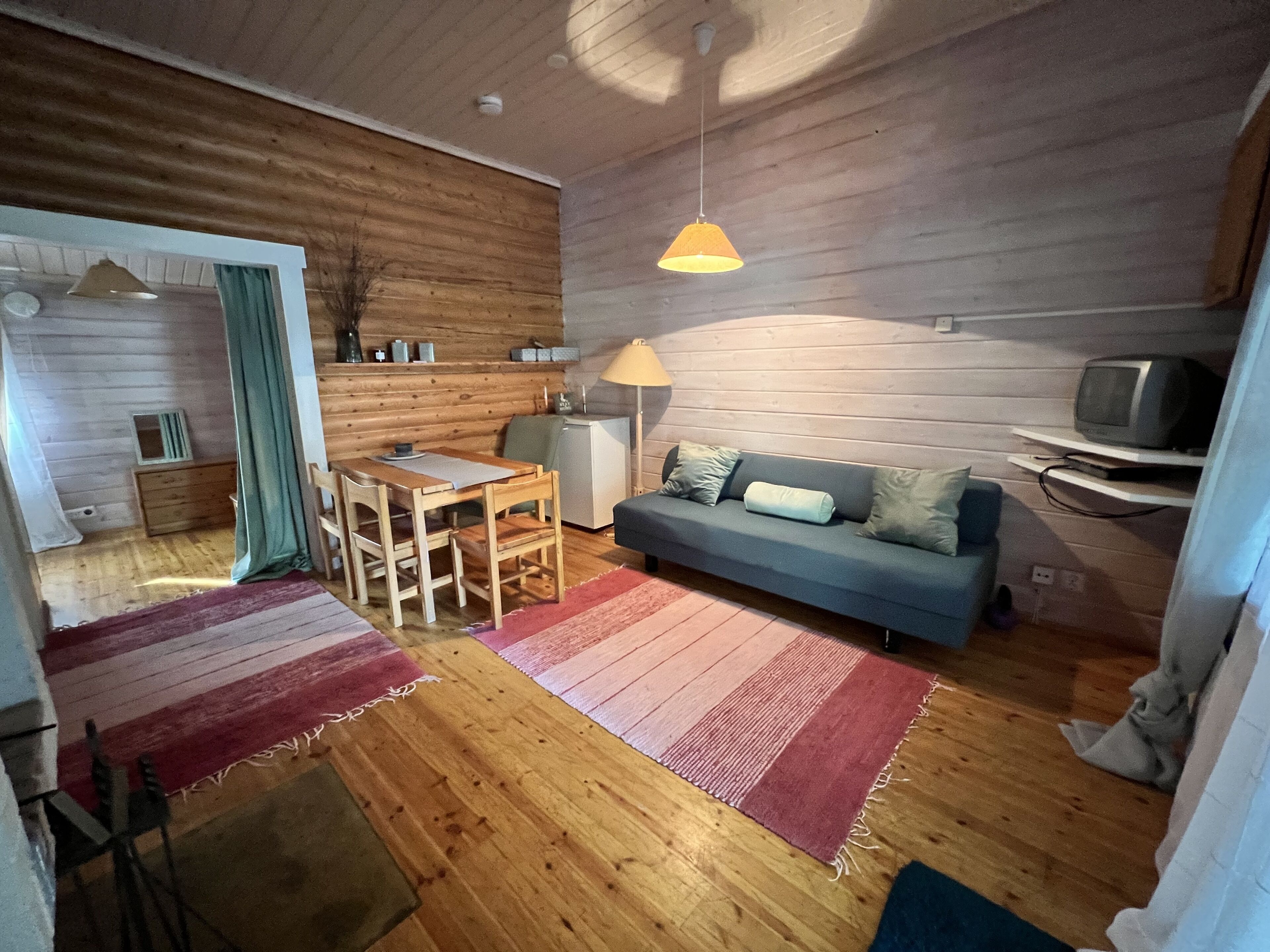 A cozy living room with wooden walls, a blue sofa, a wooden dining table with chairs, a red and pink rug, and a television on a shelf.