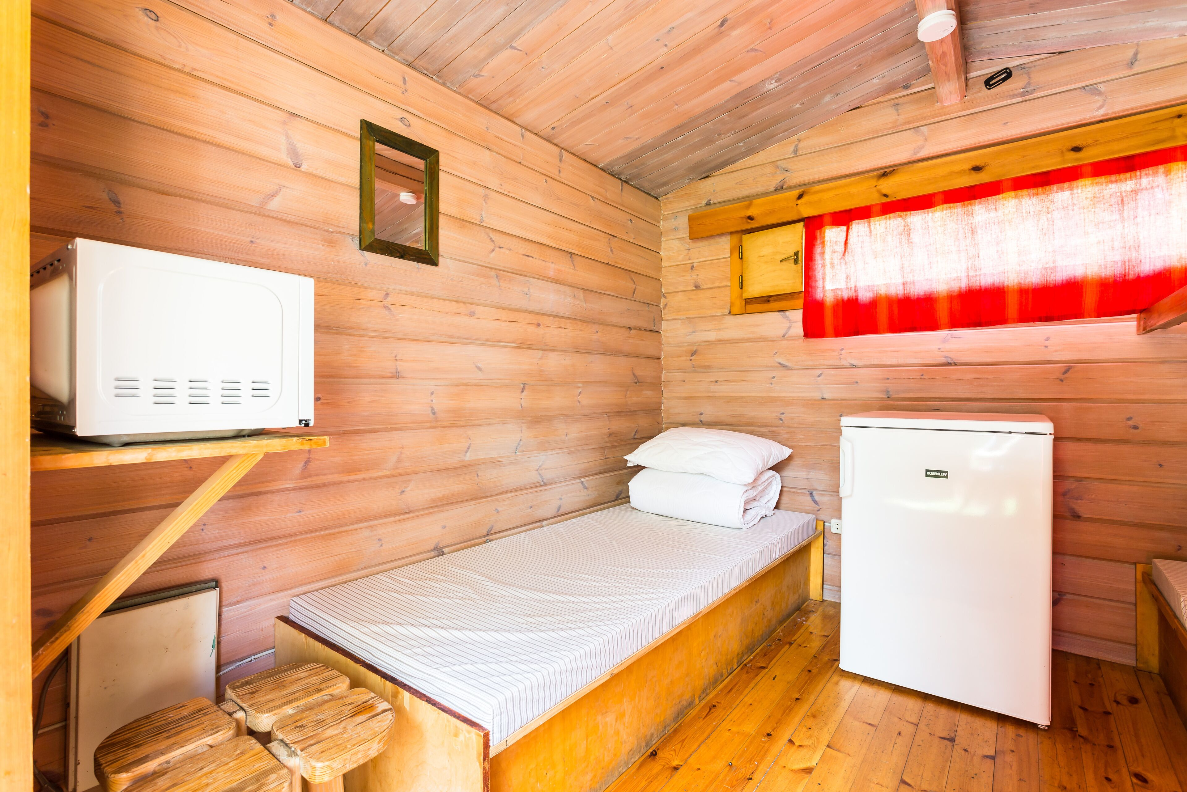 A compact wooden cabin room with a bed, a small refrigerator, a microwave, and a window with a red curtain.