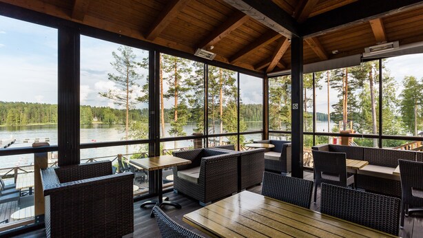 An outdoor seating area with wicker furniture and a wooden table, overlooking a lake and forest. - Punkaharju Resort (Savonlinna)
