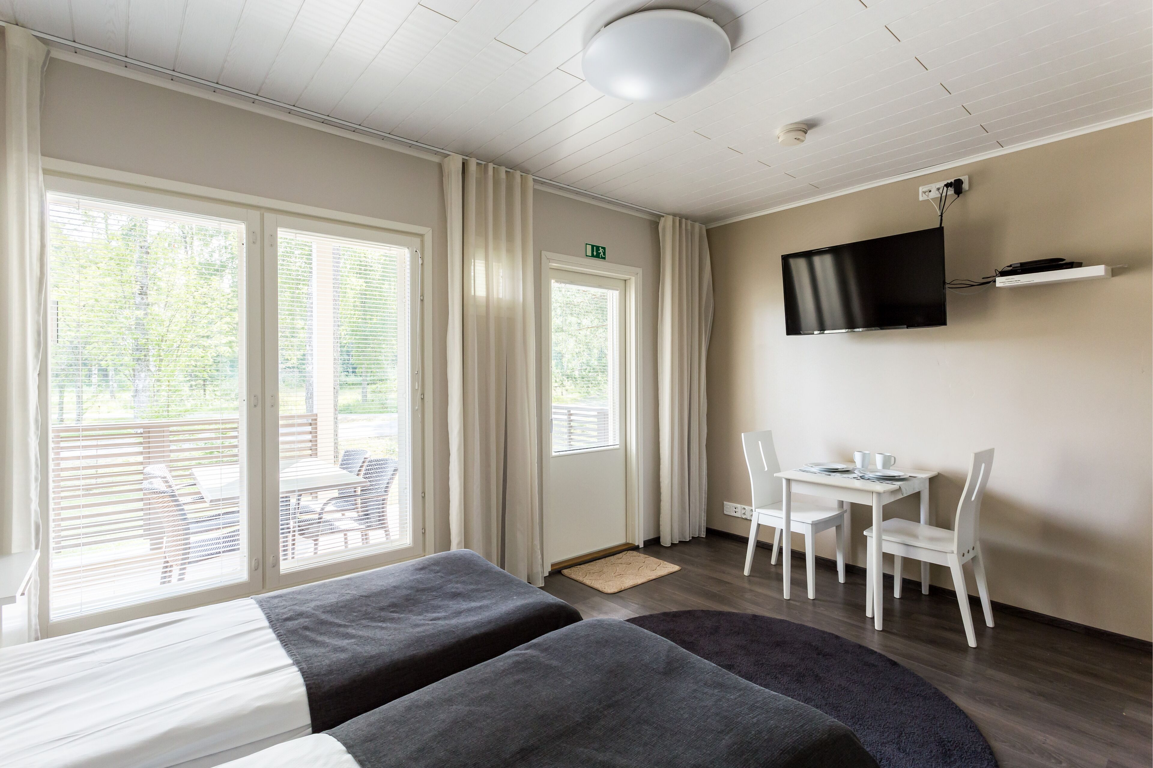 A hotel room with two beds, a TV mounted on the wall, a small table with chairs, and large windows with curtains.