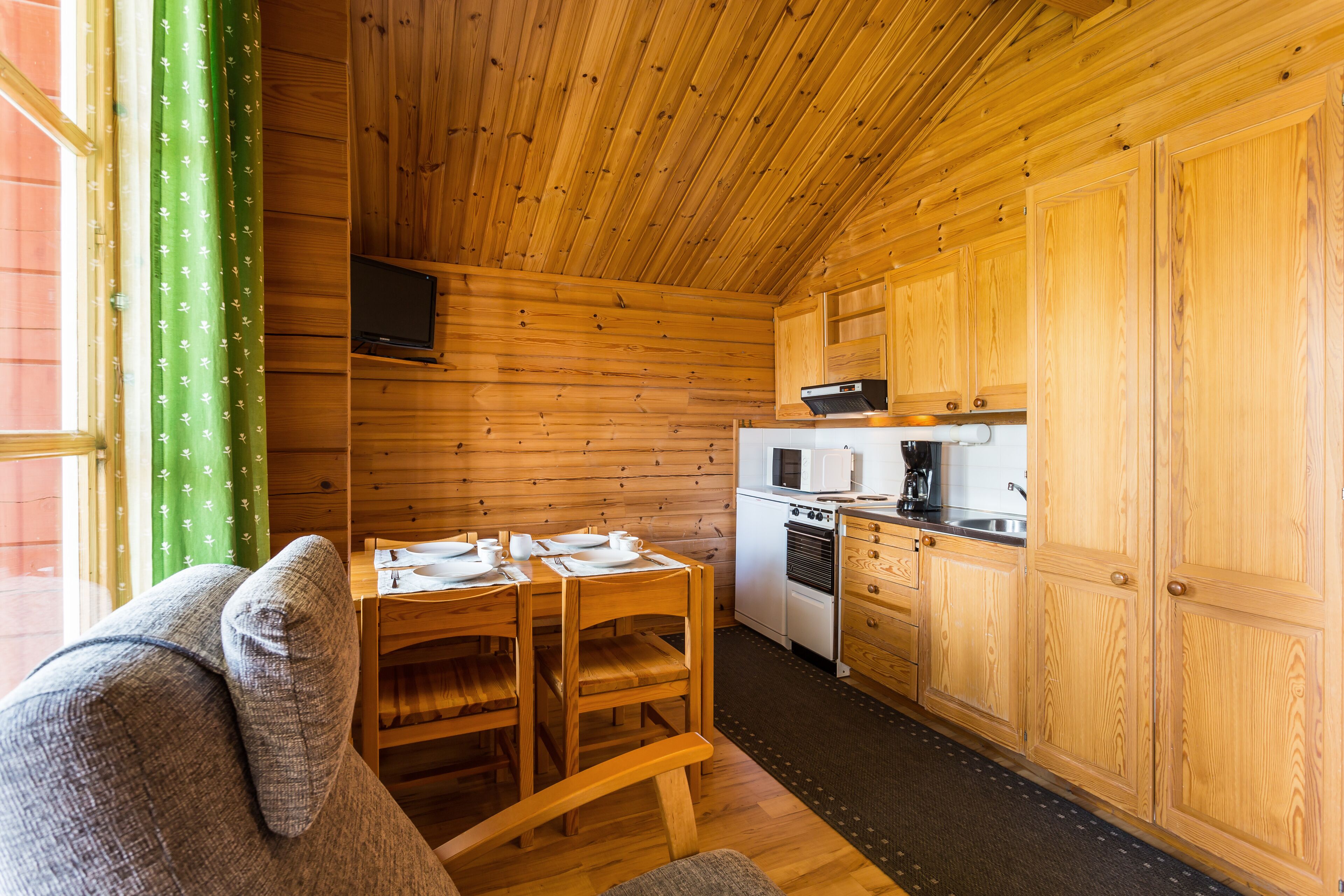 Interior de cabana de madeira com mesa de jantar posta para quatro, cozinha com micro-ondas e fogão, e cortina verde com padrão branco.