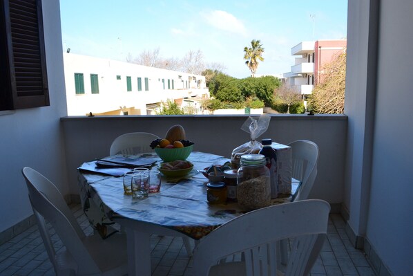 A balcony with a table set for dining, chairs, a bowl of fruit, and various food items.