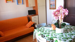 Quarto com sofá, mesa de jantar com toalha de mesa florida, vaso de flores, televisão sobre suporte e uma porta.