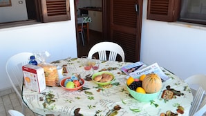 Uma mesa de jantar com diversos alimentos, uma revista e uma janela com persianas de madeira.