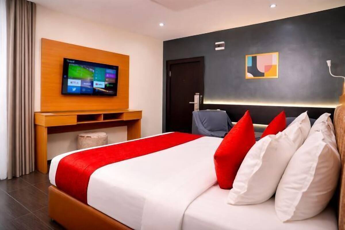 Een hotelkamer met een bed, een televisie op een houten standaard, een donkere muur met een schilderij en een deur.