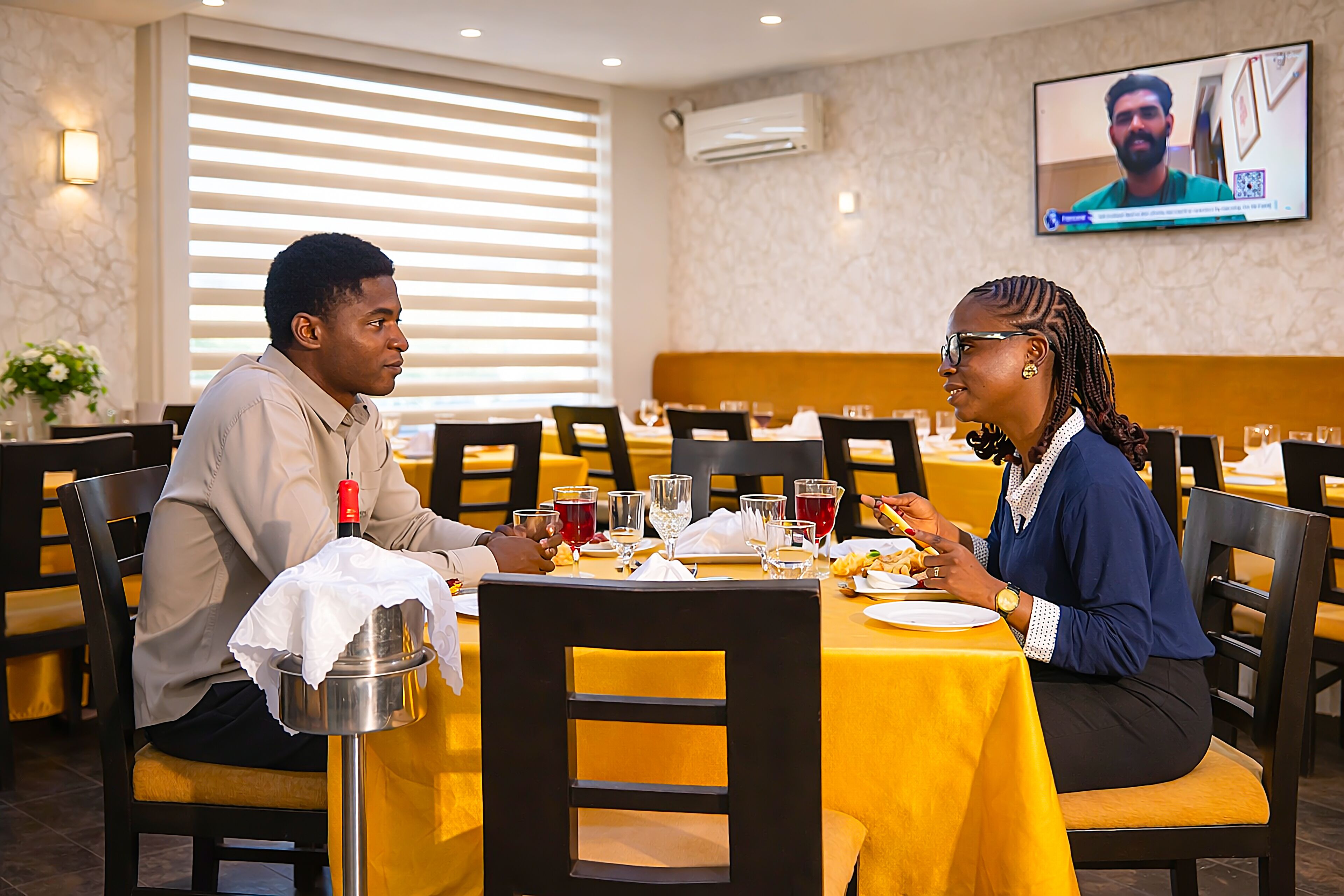 A dining area with yellow tablecloths, black chairs, and a television screen mounted on the wall.