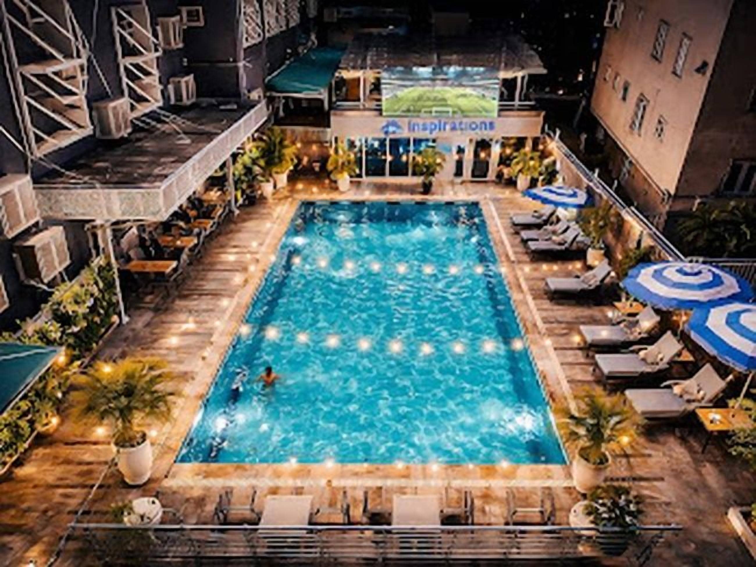 Piscina retangular com espreguiçadeiras e guarda-sóis, cercada por edifícios e área de estar ao ar livre.