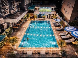 Una piscina rectangular con sillones de descanso y sombrillas, rodeada de edificios y mobiliario exterior.