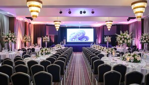 Une salle de conférence avec des rangées de chaises, des tables dressées pour un repas et un grand écran à l’avant.