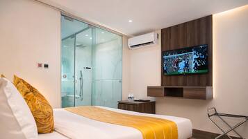 A hotel room with a bed, a television, a glass shower enclosure, and a wooden wall panel.
