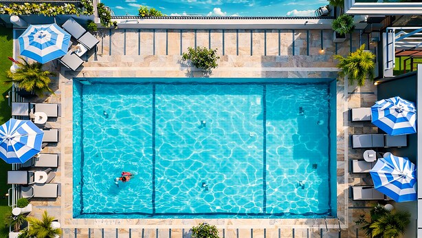 Vista aérea de una piscina rectangular con dos secciones, rodeada de sillas de descanso y sombrillas, y un muro de piedra.
