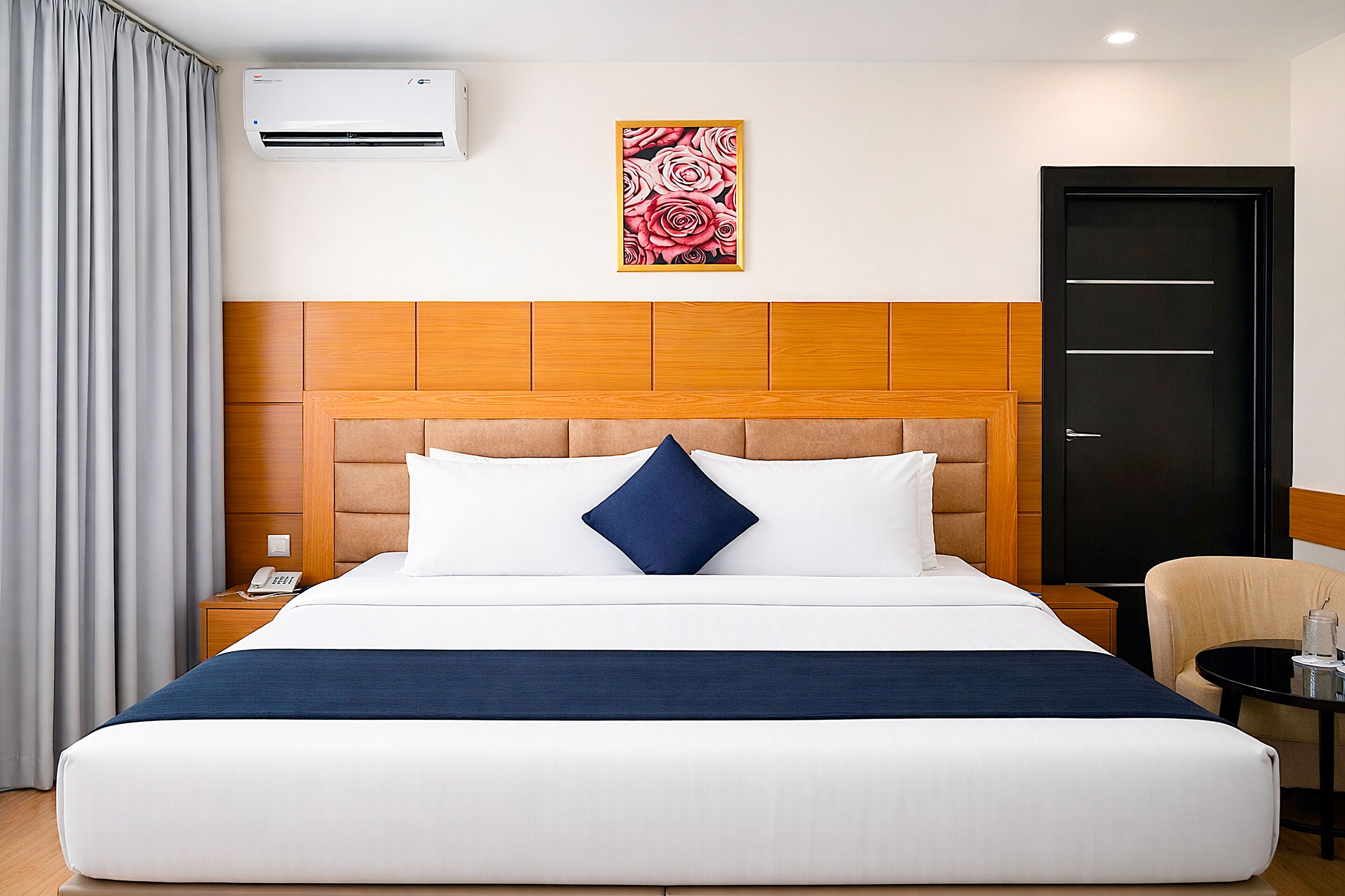 A hotel room with a large bed, a headboard, a wall-mounted air conditioner, a painting, a door, a nightstand, a chair, and a small table.