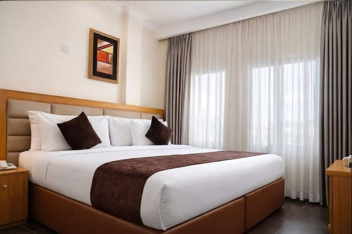 Een hotelkamer met een groot bed, een houten hoofdbord, een nachtkastje, een raam met gordijnen, en een ingelijste foto aan de muur.