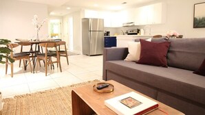 Una sala moderna con un sofá gris, una mesa de comedor de madera y sillas. Se ve una cocina al fondo con armarios blancos y un refrigerador de acero inoxidable.