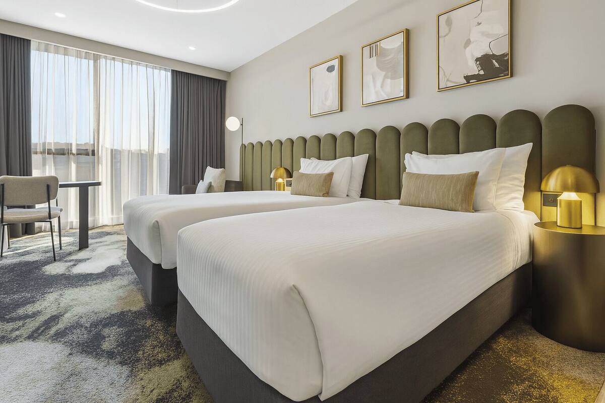 Habitación de hotel con dos camas, un escritorio y una silla. Hay cuadros en la pared y una lámpara en la mesita de noche.