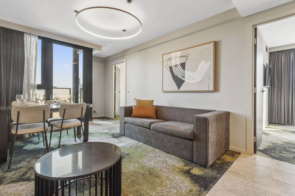 Habitación de hotel moderna con sofá, mesa de comedor y sillas. Cuenta con un ventanal con cortinas, una lámpara de techo y un cuadro en la pared.