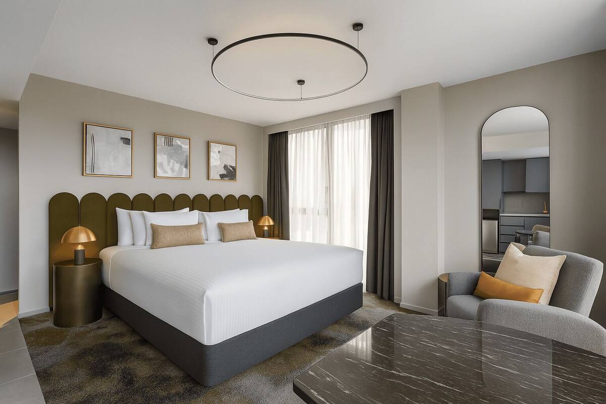 Una habitación de hotel moderna con una cama grande, una zona de estar con una mesa y vistas a la cocina.