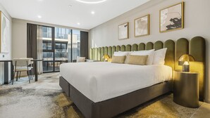 A modern hotel room with a large bed, a desk, and a view of the city through large windows.