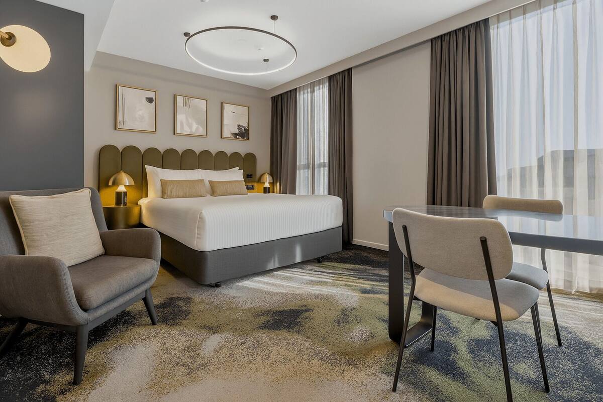 Habitación de hotel con una cama grande, dos sillones, una mesita de vidrio y vistas a la ciudad.