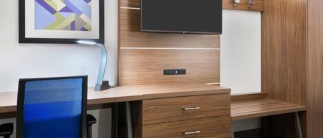 A hotel room with a wooden desk, a chair, a flat-screen TV, and a framed abstract painting on the wall.