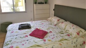 A bedroom with a floral bedspread, a green headboard, a window with a view of palm trees, and a small green chair.