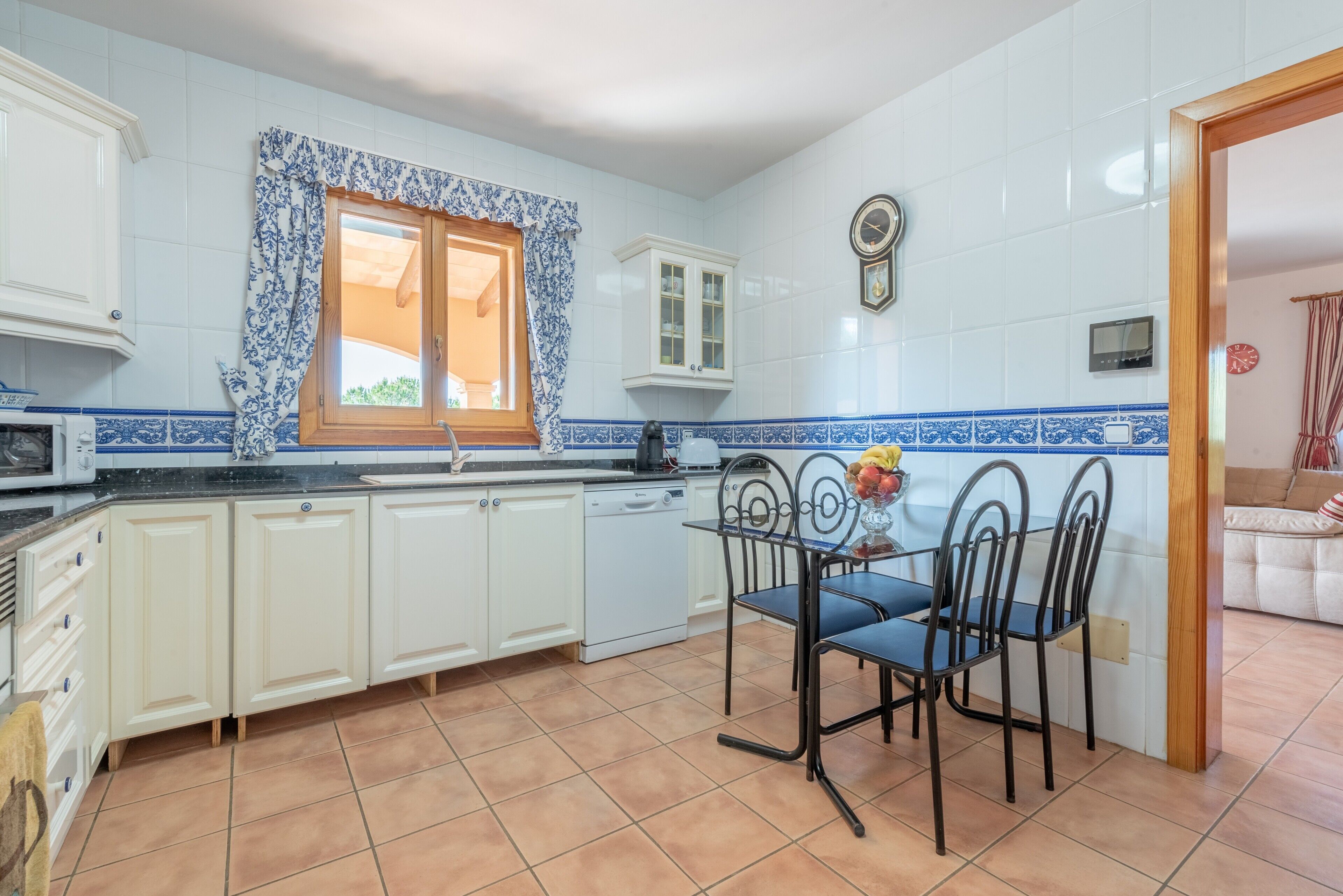 A kitchen with white cabinets, a dining table with chairs, a window with blue curtains, and a clock on the wall.