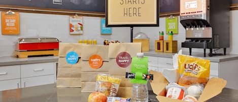 Una mesa de desayuno con un cartel que dice “HAPPY BREAKFAST STARTS HERE”, una cafetera y diversos alimentos y bolsas etiquetadas como “PURE”, “HAPPY” y “TRAVEL”.