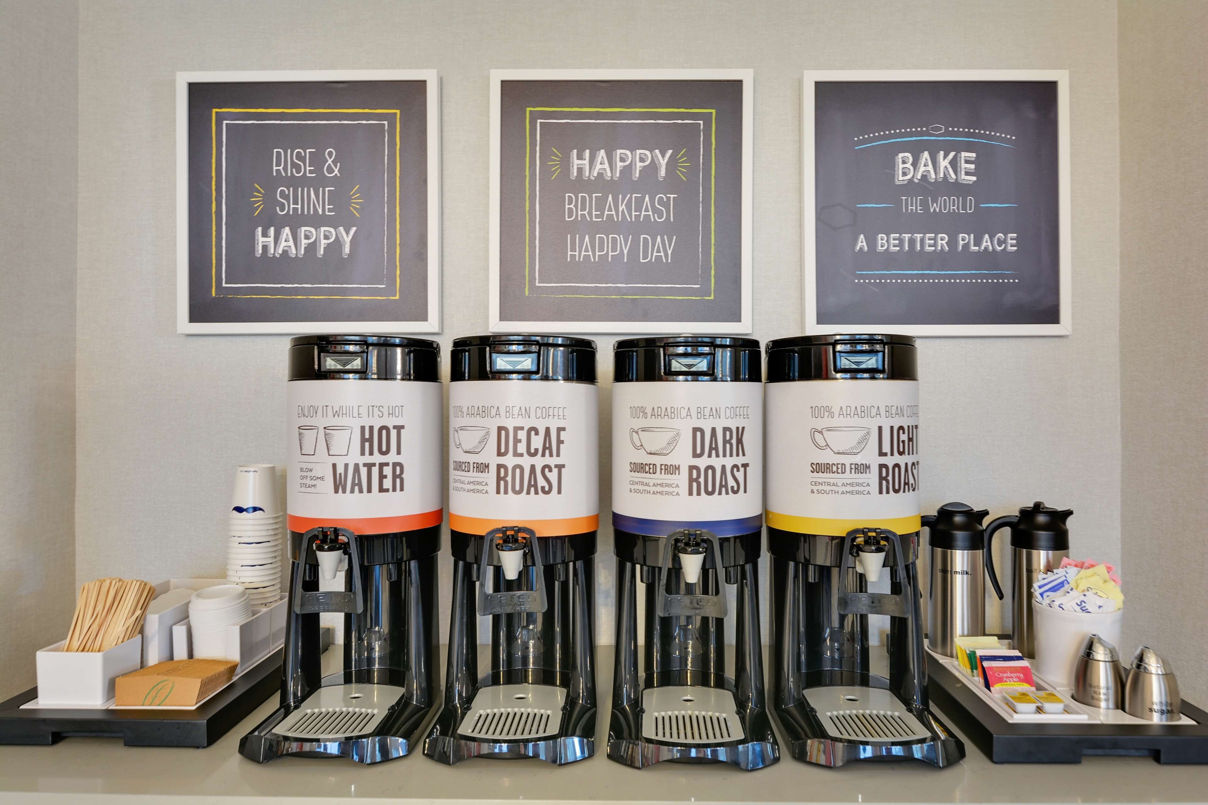 Un espace dédié au café, équipé de quatre distributeurs de café étiquetés « café décaféiné », « café torréfié foncé », « café torréfié léger » et « eau chaude ». On y trouve également des tasses à café et un plateau avec des couverts.