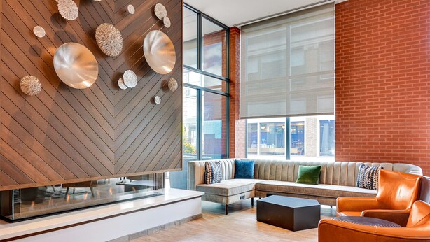 A modern interior with a herringbone wood wall, a sectional sofa with multiple cushions, a glass-top coffee table, and a brick accent wall.
