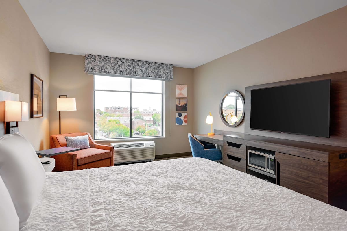 A hotel room with a large bed, a desk with a flat-screen TV, a chair, a lamp, and a window with a view of buildings.