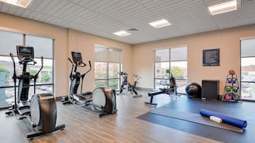 A gym with cardio machines, a yoga mat, kettlebells, and a large window with a view of buildings.