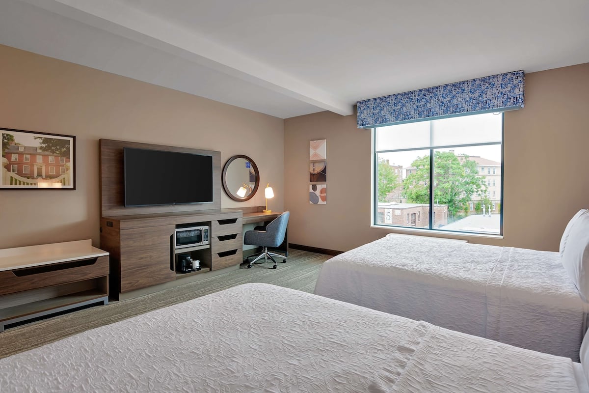 A hotel room with two beds, a TV, a desk with a chair, and a large window with a view of buildings.