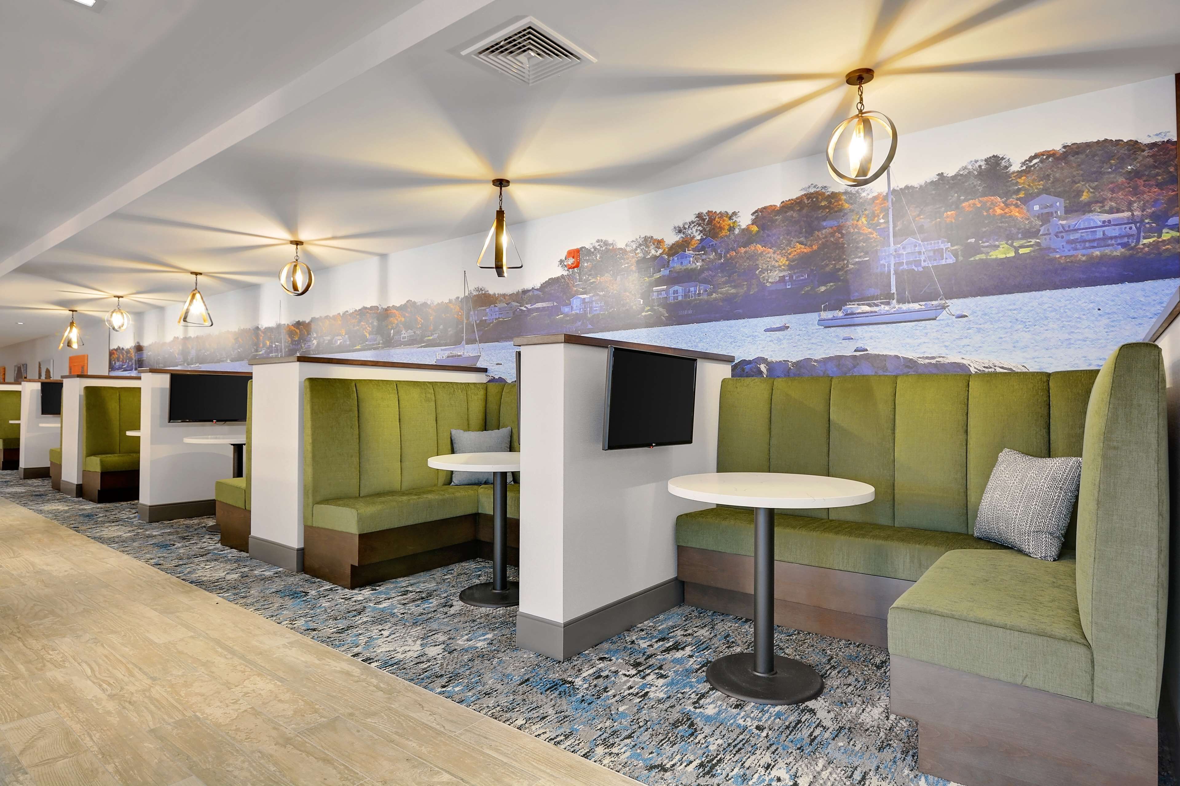 A modern hotel lobby with green seating areas, a large mural of a lakeside scene, and pendant lighting.