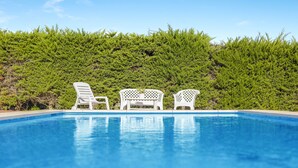 A swimming pool with three white lounge chairs and a hedge in the background.