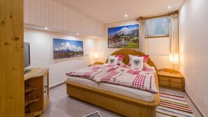 A bedroom with a wooden bed, a television on a wooden stand, two wall-mounted paintings, a window with curtains, and a bedside lamp. - Vacation Apartment Directly Between Chiemsee and The Mountains (Bernau)