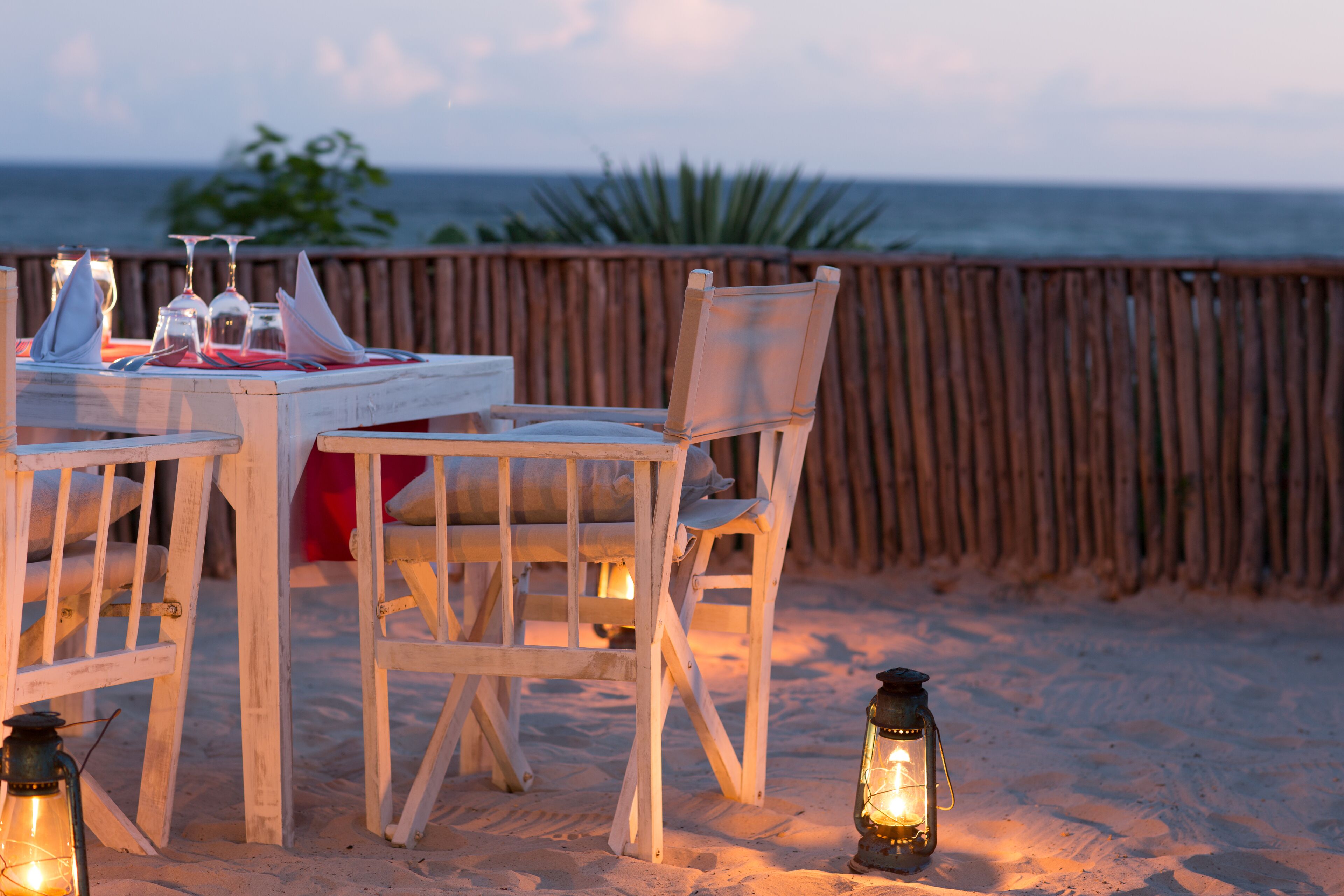 A wooden table set with glasses and a candle, surrounded by chairs, on a sandy beach with a wooden fence and ocean view.