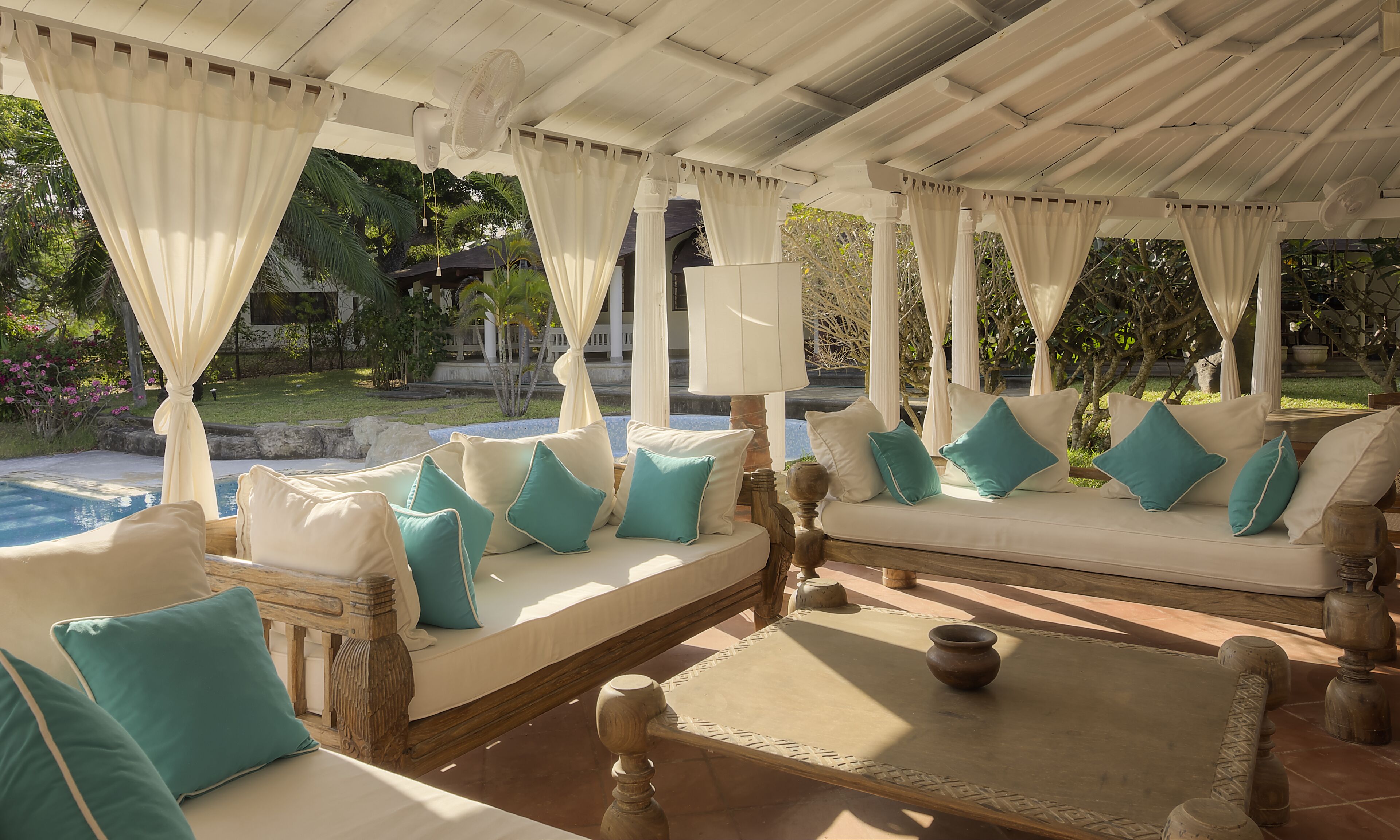 A covered outdoor seating area with white cushions and teal pillows, a wooden table, and a potted plant.
