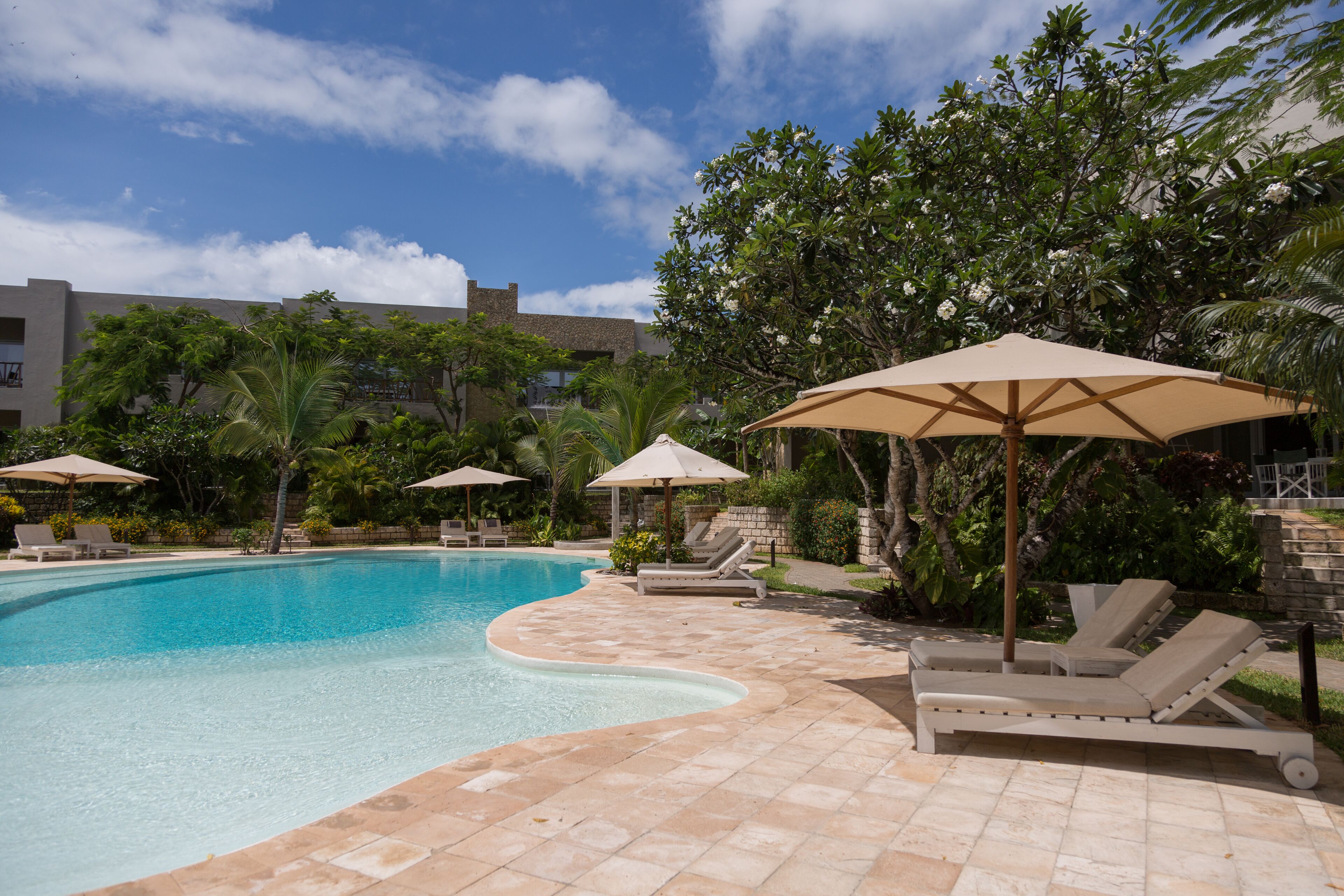 A swimming pool with lounge chairs and umbrellas, surrounded by greenery and a building in the background.
