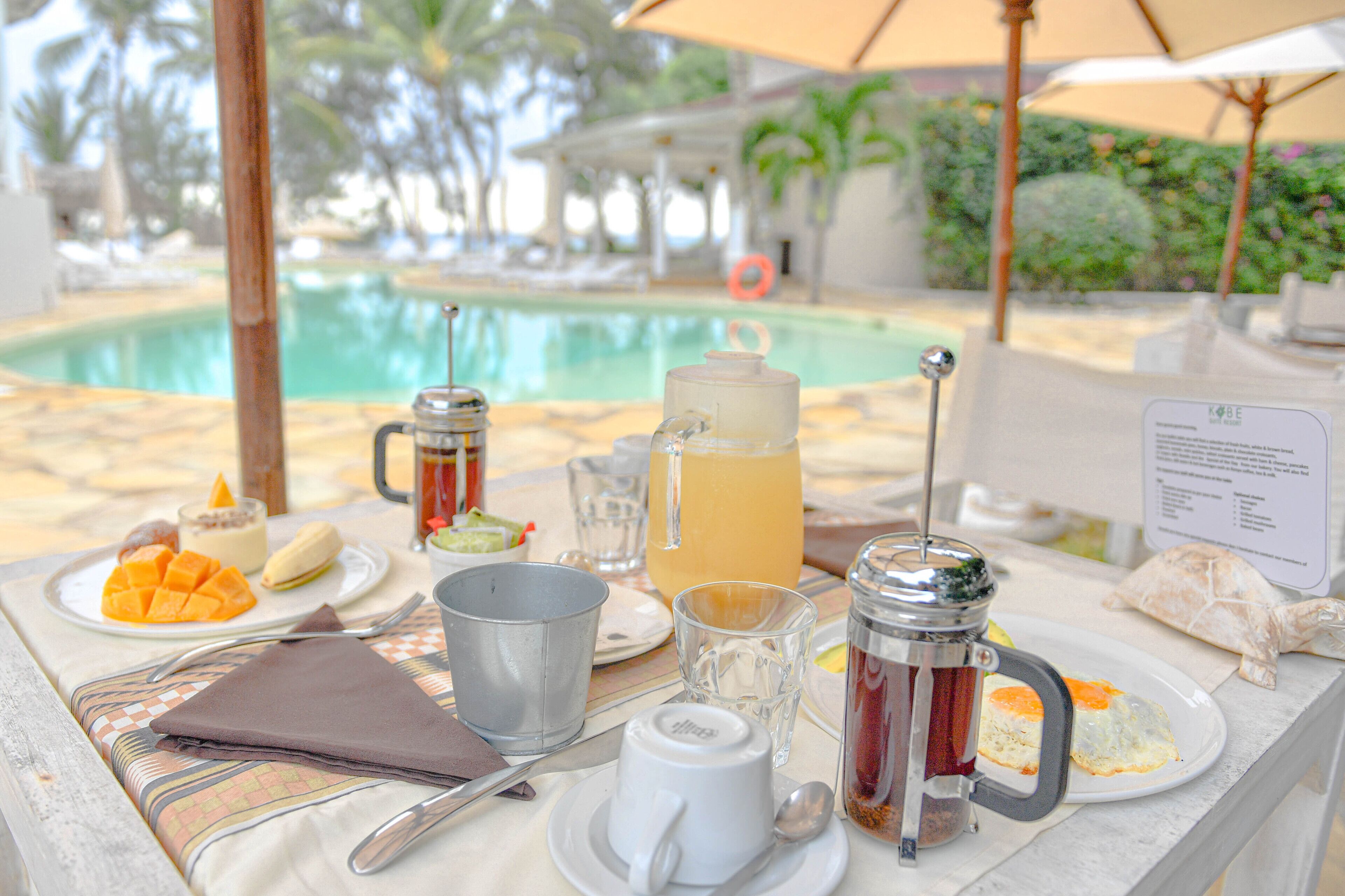 A table set with breakfast items including a fruit plate, a pitcher, a mug, and a glass of juice, with a pool and palm trees in the background.
