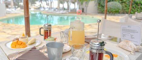 A table set with breakfast items including a fruit plate, a pitcher, a mug, and a glass of juice, with a pool and palm trees in the background.