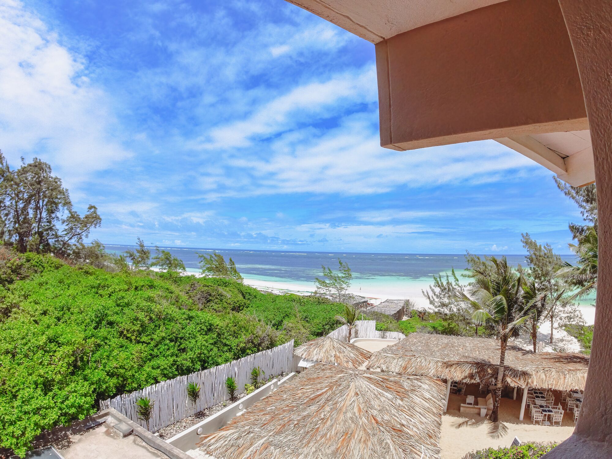 A beach view from a balcony with a thatched roof, a sandy beach, and a clear blue sky.