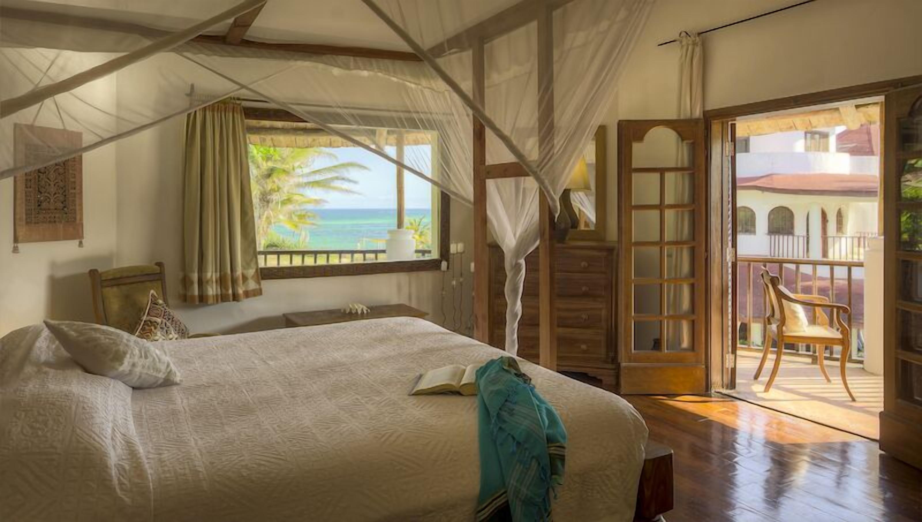 A bedroom with a large bed, a window with a view of the ocean, a wooden chair, and a balcony with a view of the ocean.