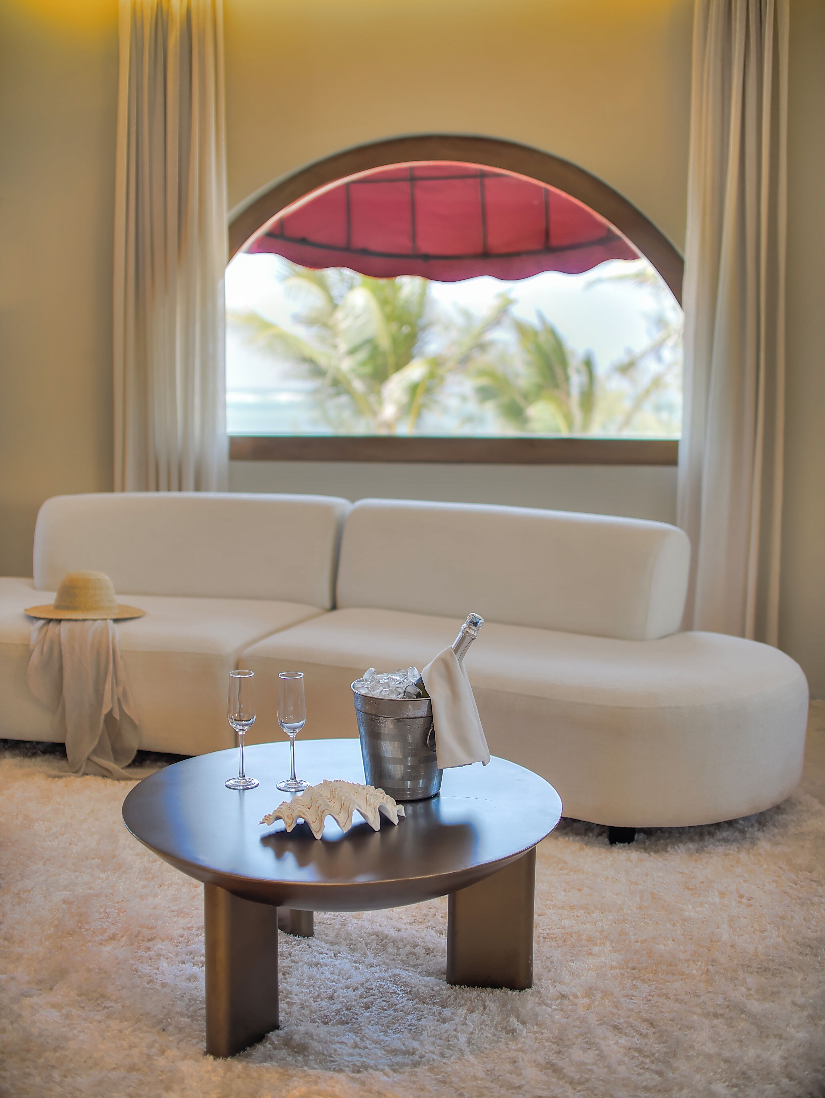 Ein weißes Sofa mit Blick auf Palmen, ein runder Tisch mit einer Champagnerflasche, einem Glas und einer dekorativen Muschel.