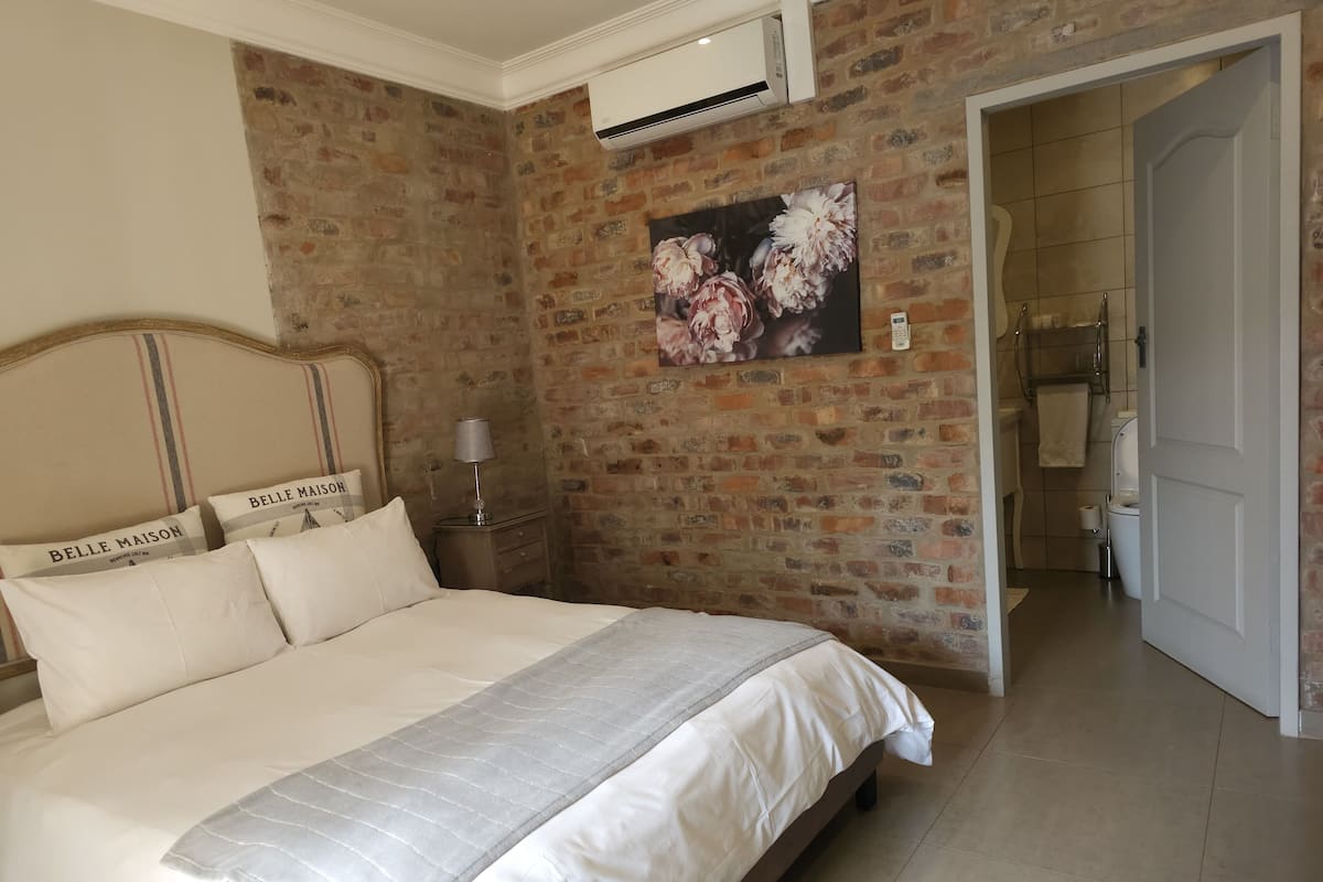 Une chambre avec un mur en briques, un lit recouvert de linge de lit blanc, une table de chevet avec une lampe, et une salle de bain visible à travers une porte ouverte.