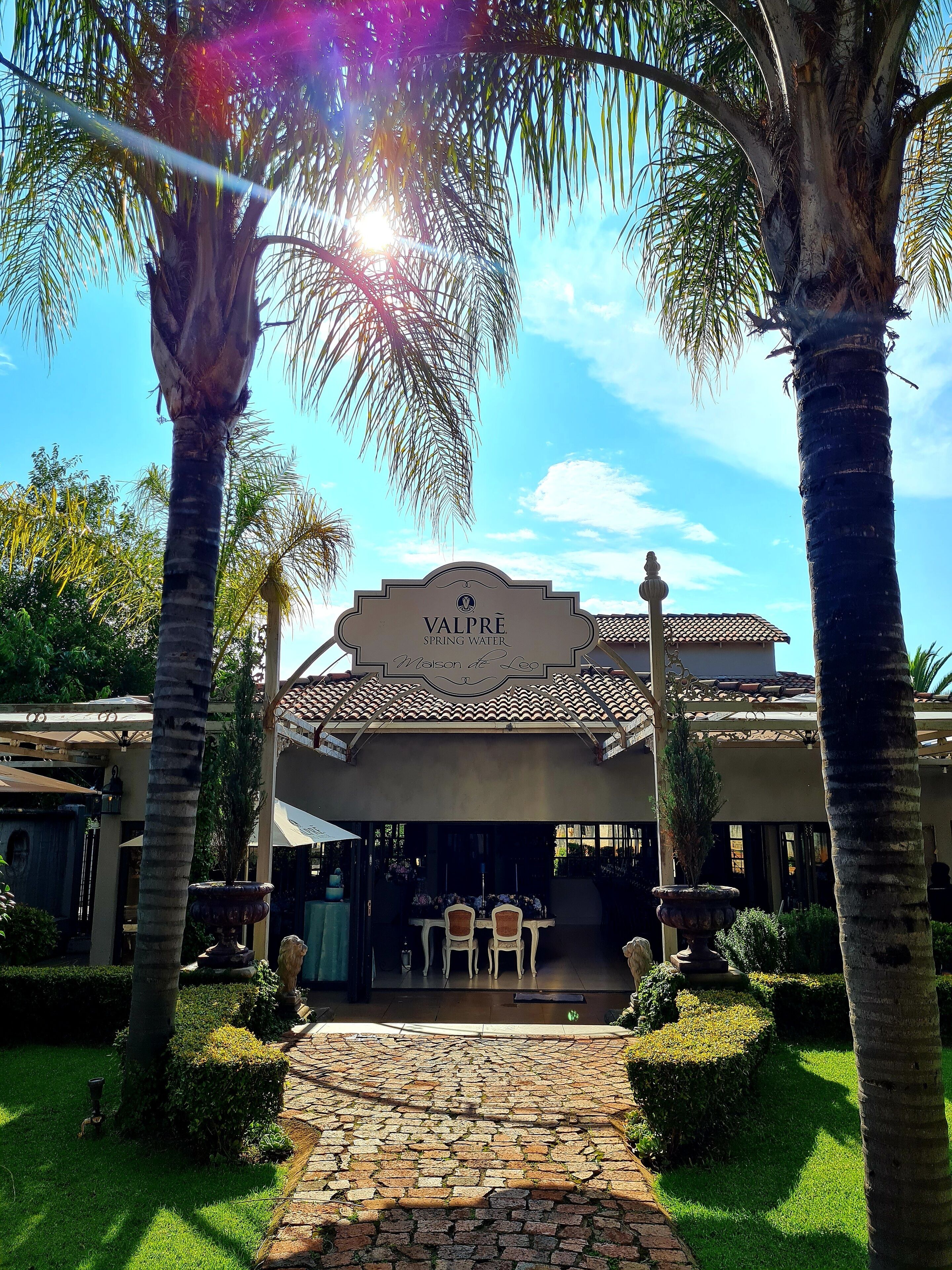 A hotel entrance with a sign reading "Valpré Spring Water." There are palm trees, a paved pathway, and a covered outdoor seating area with tables and chairs.