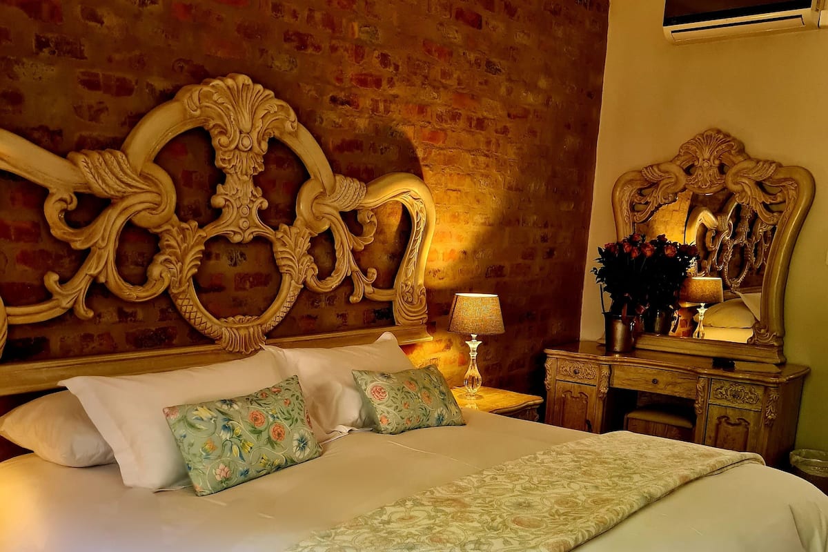 Un grand lit avec une tête de lit décorée et une literie à motifs floraux, une table de chevet avec une lampe et un miroir au-dessus.