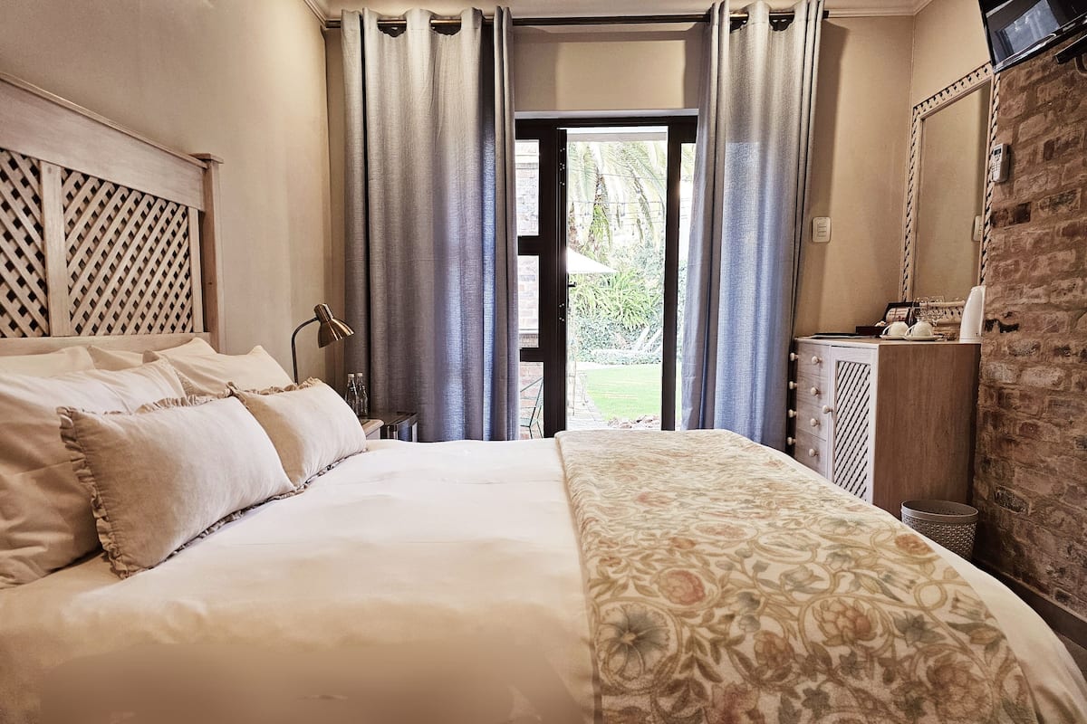 Une chambre à coucher avec un lit, des rideaux, une table de chevet, une lampe, une commode et un mur en briques.