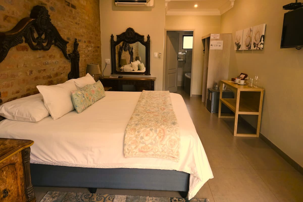 Une chambre avec un grand lit, une tête de lit en bois, un miroir, une table de chevet, une petite armoire et un téléviseur fixé au mur.