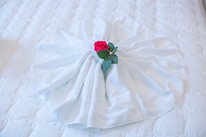 Ein ordentlich gefaltetes weißes Handtuch mit einer einzelnen roten Rose darauf, zentriert auf einem Bett mit einer gesteppten Oberfläche.