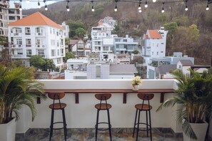 Una terraza con tres taburetes, plantas en macetas y vistas a una colina con edificios.